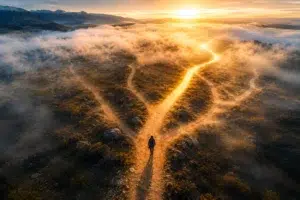 Vue aérienne d’une personne marchant sur un sentier lumineux se divisant en plusieurs chemins au lever du soleil dans un paysage brumeux.