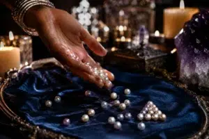 Main féminine tenant des perles blanches et rosées au-dessus d’un plateau en velours bleu nuit dans une ambiance spirituelle éclairée à la bougie.