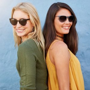 Two women smiling back to back. Stylish female friends wearing sunglasses looking at camera against blue background.